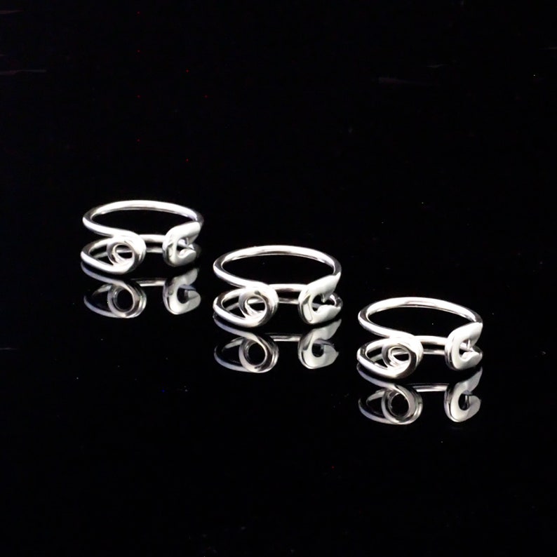 K&ouml;nnte beinhalten: Drei silberne Ringe mit einem Sicherheitsnadel-Design. Die Ringe sind auf einer schwarzen Oberfl&auml;che gestapelt.