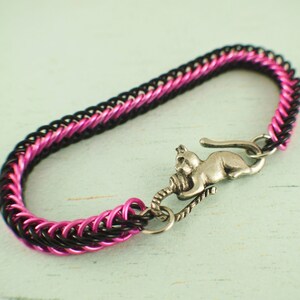 Puede incluir: Un brazalete de malla de cadena negro y rosa con un dije de gato plateado. El dije de gato sostiene una bola de lana.
