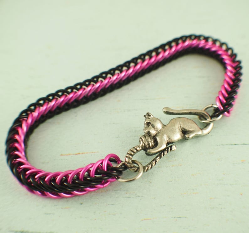 Puede incluir: Un brazalete de malla de cadena negro y rosa con un dije de gato plateado. El dije de gato sostiene una bola de lana.