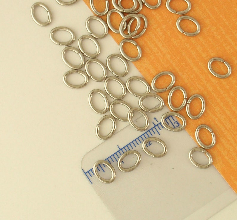 K&ouml;nnte beinhalten: Nahaufnahme eines Stapels silberner Sprungringe auf einem Lineal. Die Sprungringe sind oval geformt und werden f&uuml;r die Schmuckherstellung verwendet.