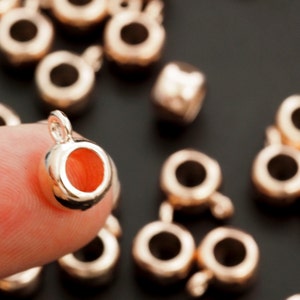May include: A close-up of a gold-toned metal jump ring with a small hole in the center. The jump ring is being held by a finger. There are other similar jump rings in the background.