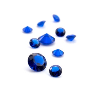 May include: A collection of ten blue gemstones, some larger than others, are scattered on a white surface. The gemstones are faceted and sparkle.