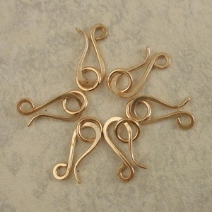 May include: Six gold-toned metal hook-and-eye jewelry clasps arranged in a circular pattern. Each clasp features a curved, S-shaped hook component and a separate circular ring component, both with a smooth, polished, and reflective metallic finish.