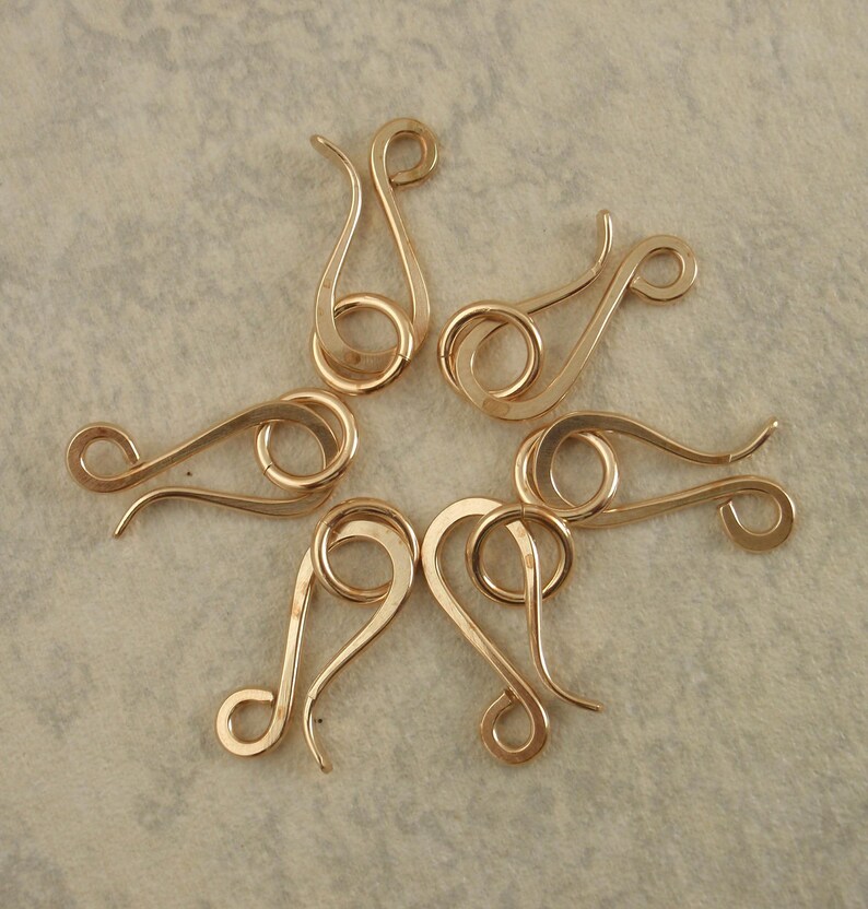 May include: Six gold-toned metal hook-and-eye jewelry clasps arranged in a circular pattern. Each clasp features a curved, S-shaped hook component and a separate circular ring component, both with a smooth, polished, and reflective metallic finish.