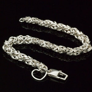 Pode incluir: Uma pulseira prateada com um design de corrente bizantina. A pulseira apresenta um berloque em forma de coração e um fecho retangular. A corrente é feita de elos circulares interconectados. A pulseira é exibida em um fundo preto.