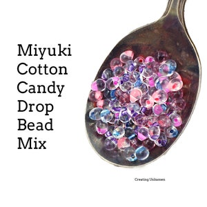 May include: A silver spoon filled with a mix of clear, pink, purple, and blue Miyuki Cotton Candy Drop Beads.