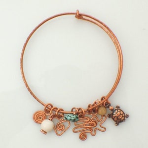 May include: A copper wire bangle bracelet featuring a collection of charms including a small copper turtle, a smooth white bead, a tan bead, a turquoise-coloured stone, and several copper wire shapes including spirals and a zigzag pattern.