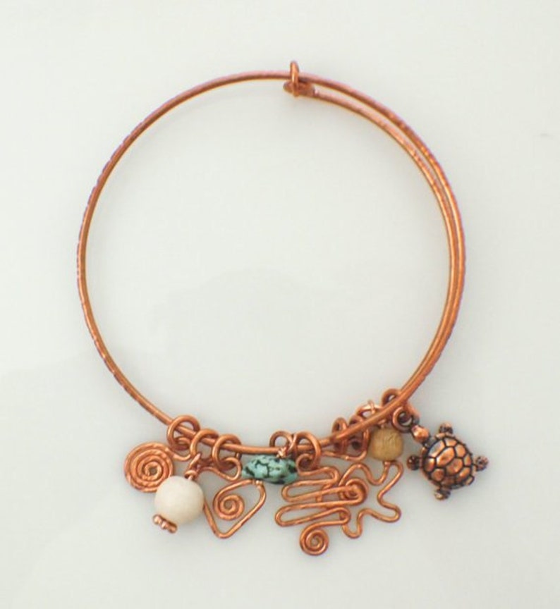 May include: A copper wire bangle bracelet featuring a collection of charms including a small copper turtle, a smooth white bead, a tan bead, a turquoise-coloured stone, and several copper wire shapes including spirals and a zigzag pattern.