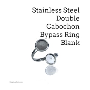 May include: A stainless steel double cabochon bypass ring blank. The ring has two circular settings and a curved band. The text "Stainless Steel Double Cabochon Bypass Ring Blank" is displayed in black. The ring is shown on a white background.