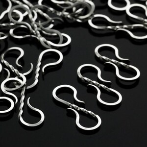 May include: Close-up of silver-colored S-shaped clasps, scattered on a dark surface. The clasps have a twisted design on the central bar and are likely used for jewelry making or crafts.