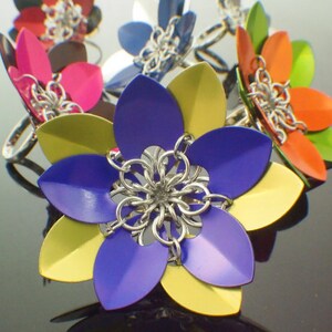 May include: A colorful set of seven metal flower rings. The rings are made of chainmail and have a variety of colors, including blue, yellow, green, orange, and pink.