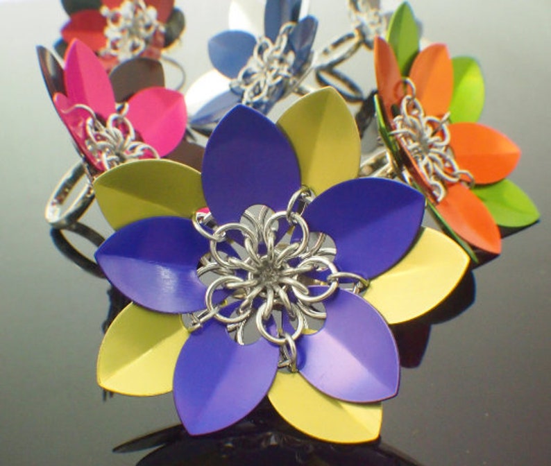 May include: A colorful set of seven metal flower rings. The rings are made of chainmail and have a variety of colors, including blue, yellow, green, orange, and pink.