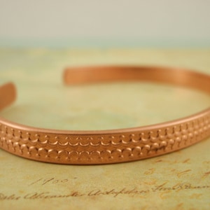 May include: A copper bracelet with a textured design. The bracelet is a simple, solid band with a repeating pattern of small circles.