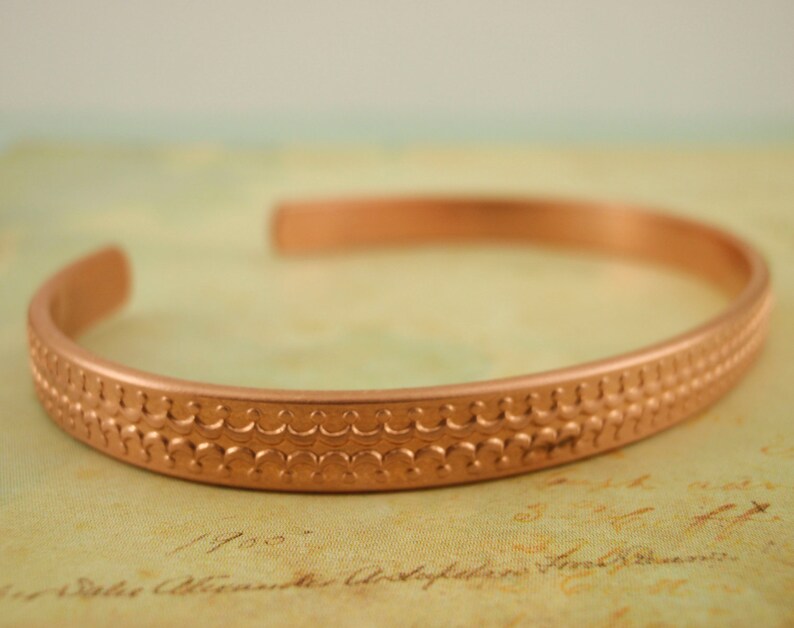 May include: A copper bracelet with a textured design. The bracelet is a simple, solid band with a repeating pattern of small circles.