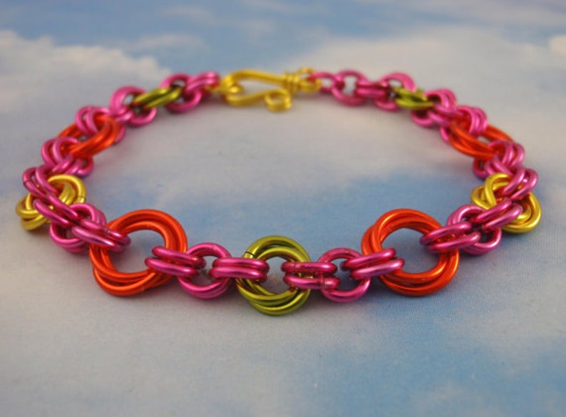 K&ouml;nnte beinhalten: Ein Kettenarmband aus ineinandergreifenden Metallringen in leuchtenden Farben wie Pink, leuchtendem Orange und Limettengr&uuml;n. Das Armband verf&uuml;gt &uuml;ber einen goldfarbenen S-Haken-Verschluss, der die kreisf&ouml;rmigen Glieder zu einer Schlaufe verbindet.