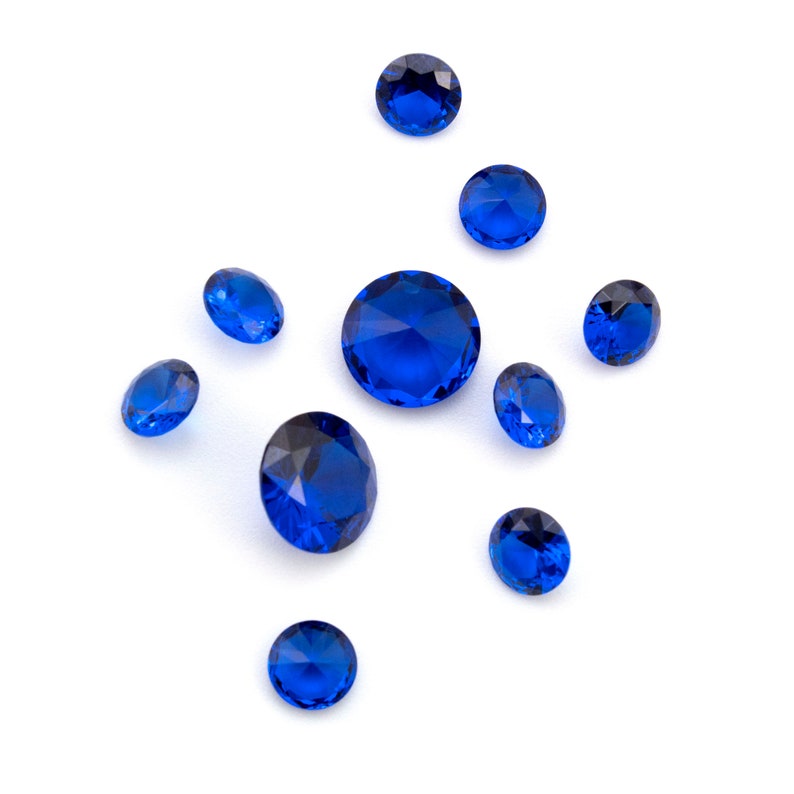 May include: A scattering of ten blue gemstones on a white background. The gemstones are various sizes and shapes, with some being round and others being more elongated.