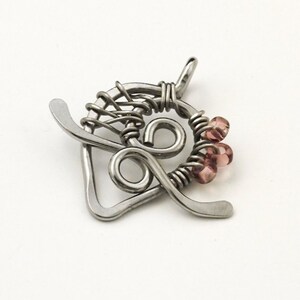 May include: A silver-toned wire-wrapped pendant featuring a heart-shaped frame with a central spiral design. The metal wire is coiled around the frame and accented with three small, translucent, light pink beads on the right side.