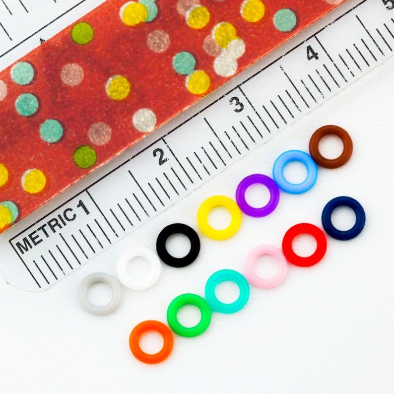 K&ouml;nnte beinhalten: Ein Satz von 10 bunten Gummiringen, die in einer Reihe auf einer wei&szlig;en Oberfl&auml;che angeordnet sind. Die Ringe sind in verschiedenen Farben, darunter rot, orange, gelb, gr&uuml;n, blau, lila, rosa, wei&szlig;, grau und schwarz.