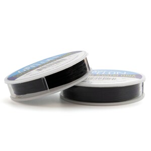 May include: Two round plastic spools of black jewelry fiber, labeled Offelon Stretch Jewelry Fiber. The spools are stacked, showing a thin, dark, flexible cord wound tightly around the center. Each spool features a blue and white label with text.