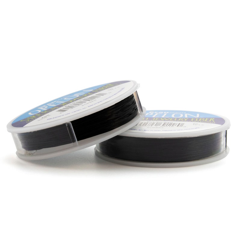 May include: Two round plastic spools of black jewelry fiber, labeled Offelon Stretch Jewelry Fiber. The spools are stacked, showing a thin, dark, flexible cord wound tightly around the center. Each spool features a blue and white label with text.