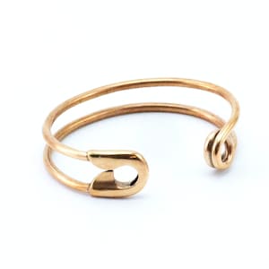 K&ouml;nnte beinhalten: Ein goldfarbener Sicherheitsnadelring. Der Ring besteht aus zwei Metallb&auml;ndern, die oben an der Sicherheitsnadel zusammengef&uuml;gt sind. Der Ring ist hinten offen, so dass er an verschiedene Fingergr&ouml;&szlig;en angepasst werden kann.