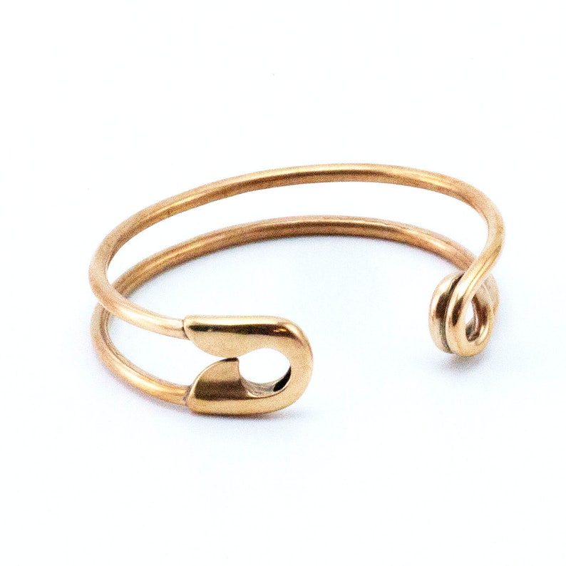 K&ouml;nnte beinhalten: Ein goldfarbener Sicherheitsnadelring. Der Ring besteht aus zwei Metallb&auml;ndern, die oben an der Sicherheitsnadel zusammengef&uuml;gt sind. Der Ring ist hinten offen, so dass er an verschiedene Fingergr&ouml;&szlig;en angepasst werden kann.