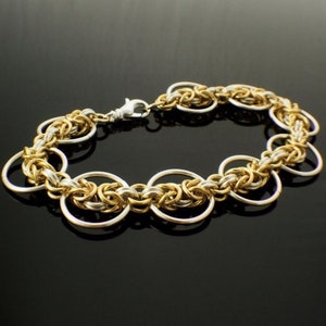 May include: A chainmail bracelet constructed from interlocking gold and silver-toned metal rings. The design features a repeating pattern of smaller woven links connected to larger, smooth circular rings, secured with a silver-toned lobster clasp.