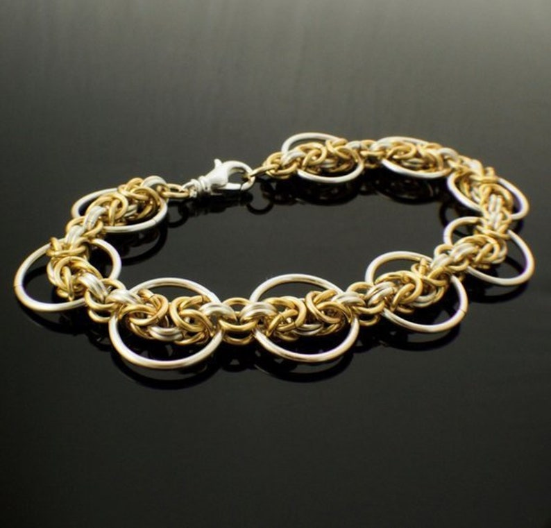 May include: A chainmail bracelet constructed from interlocking gold and silver-toned metal rings. The design features a repeating pattern of smaller woven links connected to larger, smooth circular rings, secured with a silver-toned lobster clasp.