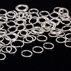 May include: A large pile of silver jump rings, scattered on a black surface. The rings are all the same size and are made of metal.