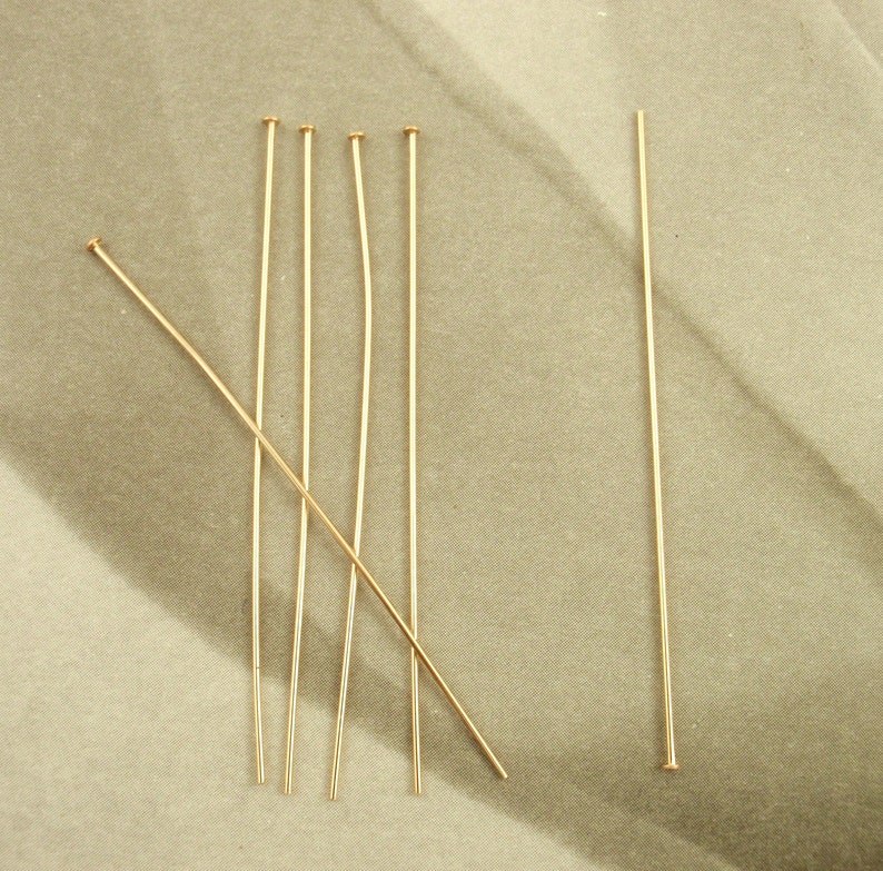 Puede incluir: Un conjunto de seis alfileres de cabeza de metal dorado, finos y delgados, cada uno con una peque&ntilde;a cabeza circular plana en un extremo y un v&aacute;stago recto. Cinco alfileres est&aacute;n dispuestos verticalmente en grupo y uno en diagonal.