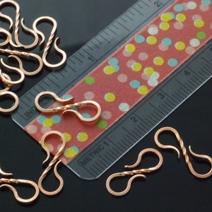 May include: Copper-colored S-shaped jewelry clasps, some with a twisted design, are scattered across a black surface. A clear ruler with inch markings and a red tape measure with colorful polka dots are also present, providing scale.