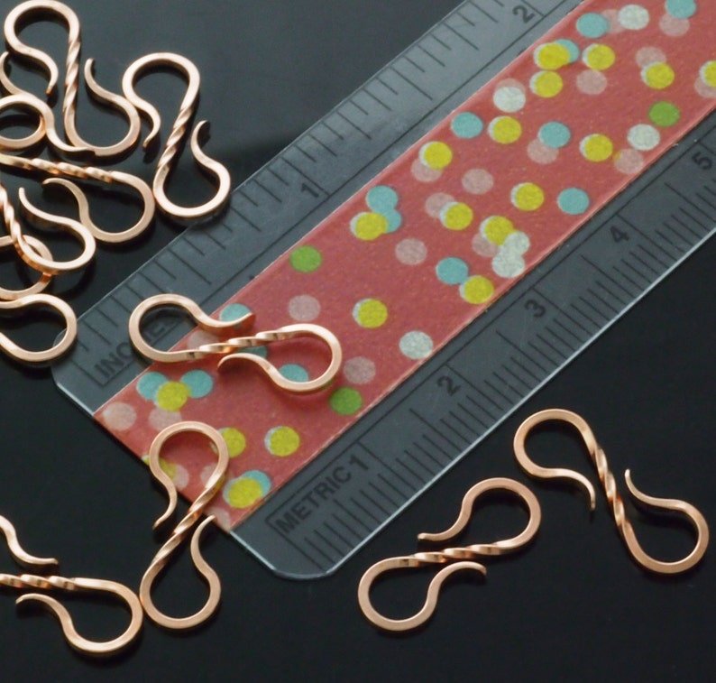 May include: Copper-colored S-shaped jewelry clasps, some with a twisted design, are scattered across a black surface. A clear ruler with inch markings and a red tape measure with colorful polka dots are also present, providing scale.