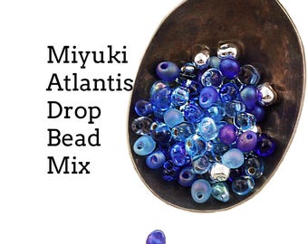 Może przedstawiać: Mieszanka koralików łezek Miyuki Atlantis w odcieniach niebieskiego, fioletowego i srebrnego, umieszczona w ciemnej metalowej łyżce. Tekst "Miyuki Atlantis Drop Bead Mix" jest wydrukowany po lewej stronie, a "Creating Unkamen" na dole.