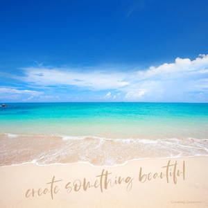 May include: A beach scene with blue sky, white clouds, and turquoise water. The sand is white and the waves are lapping at the shore. The text "create something beautiful" is written on the sand.