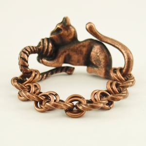 Puede incluir: Un anillo de cobre con un dise&ntilde;o de cuerda trenzada y una peque&ntilde;a figura de gato sosteniendo una bola de lana. El gato est&aacute; de espaldas al espectador y su cola est&aacute; enroscada alrededor del anillo.