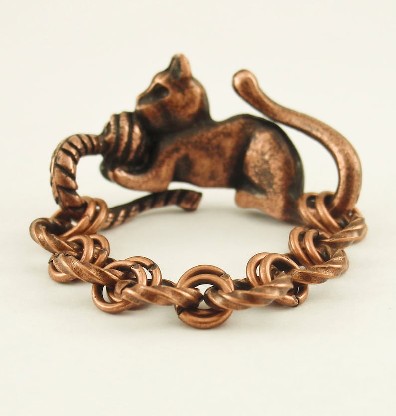 Puede incluir: Un anillo de cobre con un dise&ntilde;o de cuerda trenzada y una peque&ntilde;a figura de gato sosteniendo una bola de lana. El gato est&aacute; de espaldas al espectador y su cola est&aacute; enroscada alrededor del anillo.