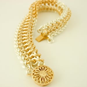 May include: A chainmail bracelet featuring alternating silver and gold-toned metal rings in a woven pattern. The bracelet includes a round, gold-toned decorative clasp with a radial, flower-like cutout design and a rectangular metal fastener.