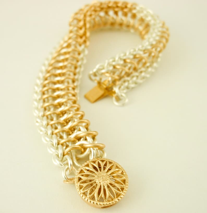 May include: A chainmail bracelet featuring alternating silver and gold-toned metal rings in a woven pattern. The bracelet includes a round, gold-toned decorative clasp with a radial, flower-like cutout design and a rectangular metal fastener.