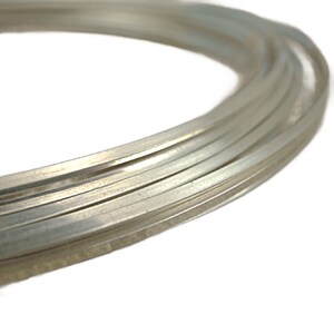 May include: A continuous coil of flat, silver-colored metal wire. The wire has a smooth, reflective surface and is wound into several overlapping circular loops, showcasing a consistent rectangular cross-section throughout the entire length.