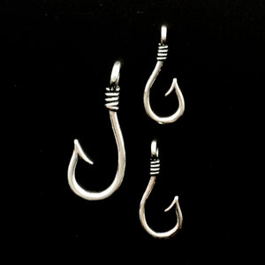 May include: Three silver-coloured fish hook pendants against a black background. The hooks vary in size, with a wrapped wire detail at the top. The hooks are curved with a sharp point, suggesting a fishing or nautical theme.