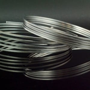May include: A coiled length of flat, silver-colored metal wire. The wire is wound into multiple overlapping circular loops, creating a layered, spiral appearance. The metallic surface has a smooth, reflective finish with visible edges.
