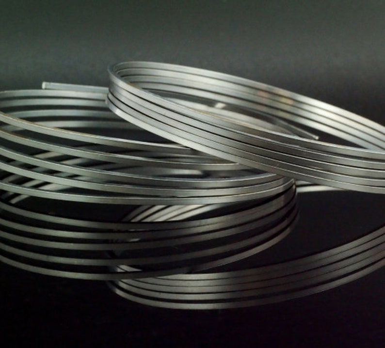 May include: A coiled length of flat, silver-colored metal wire. The wire is wound into multiple overlapping circular loops, creating a layered, spiral appearance. The metallic surface has a smooth, reflective finish with visible edges.