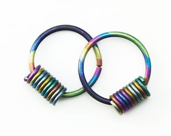 Puede incluir: Dos pendientes de aro iridiscentes con un diseño en espiral. Los pendientes presentan un espectro de colores, incluyendo azules, verdes, morados y amarillos. Los aros están conectados por una pequeña sección articulada. Sobre fondo blanco.