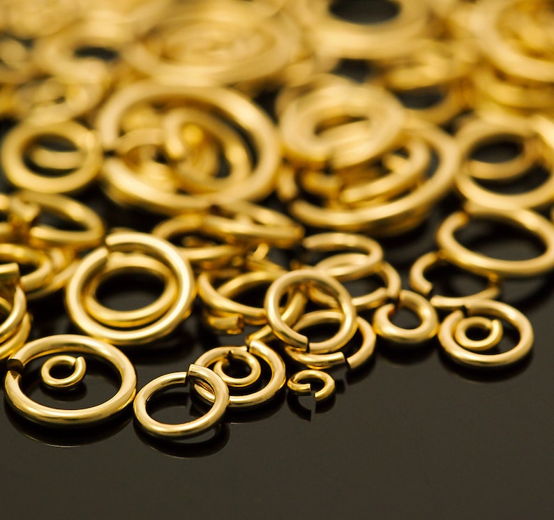 Op de afbeelding: Een close-up van talrijke goudkleurige verbindingsringen in verschillende maten, verspreid over een donkere ondergrond. Sommige ringen zijn open, andere gesloten. De belichting benadrukt de metallic glans van deze sieradenbenodigdheden.