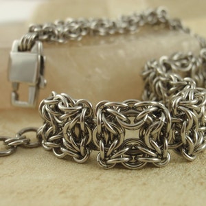 May include: A silver-toned bracelet with a woven design. The bracelet features a series of square, interconnected links, and a chain. The clasp is a simple, rectangular design. The jewelry is displayed on a neutral background.
