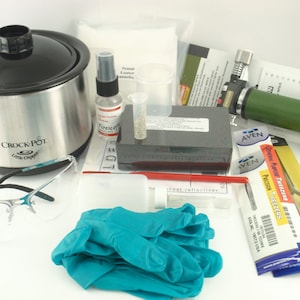 May include: A collection of jewelry-making tools and supplies, including a Crock-Pot, safety glasses, a spray bottle, tweezers, gloves, and a small torch. The items are arranged on a white surface.