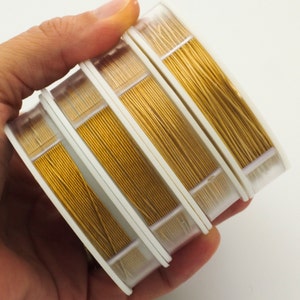 May include: Four circular spools of metallic gold-colored craft wire, each featuring a distinct thickness. The spools are stacked and held together, showcasing the shiny, reflective surface of the thin wire wound neatly around white plastic cores.