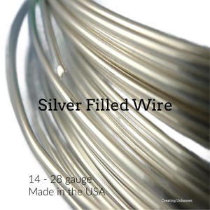 Puede incluir: Una bobina de alambre relleno de plata con un acabado metálico liso. El texto en la imagen dice: Silver Filled Wire, 14 - 28 gauge, Made in the USA y Creating Unkamen. El alambre está dispuesto en múltiples bucles circulares.
