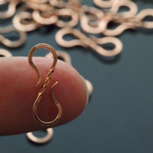 May include: Close-up of a copper-colored hook clasp with a twisted center, held between fingers. The clasp has a curved top and bottom, with several similar clasps out of focus in the background. The background is a dark teal color.