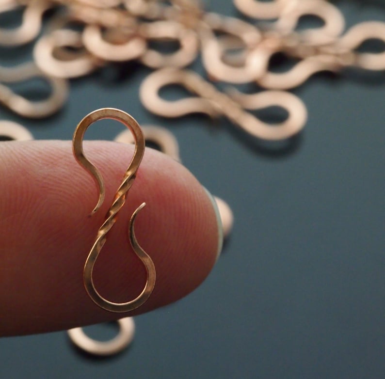 May include: Close-up of a copper-colored hook clasp with a twisted center, held between fingers. The clasp has a curved top and bottom, with several similar clasps out of focus in the background. The background is a dark teal color.